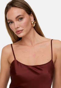 Burgunderrotes Satin-Top mit dünnen Trägern und eckigem Ausschnitt, kombiniert mit auffälligen goldfarbenen Spiralohrringen. Glatte Textur mit Glanz.