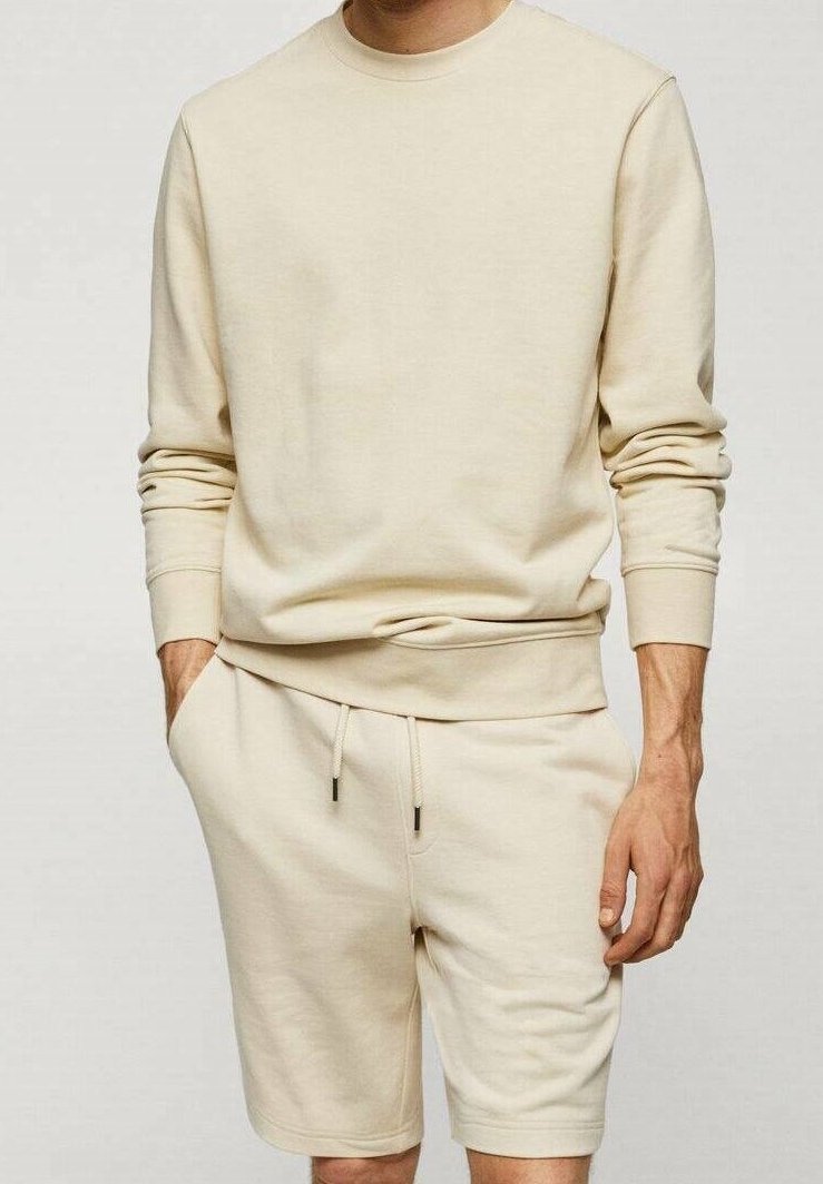 Homme portant un sweat-shirt beige clair à manches longues et un short à cordon assorti, debout avec une main dans la poche.