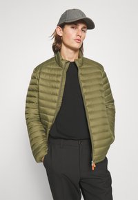 Olijfgroene pufferjack met horizontale stiksels, opstaande kraag en rits. Draag het over een zwart shirt en gecombineerd met donkere broeken. Grijze pet.