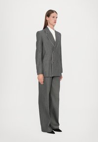Grå dobbeltradet blazer med hvit piping, sammen med brede grå bukser. Stoffet har en teksturert overflate, og svarte sko fullfører antrekket.