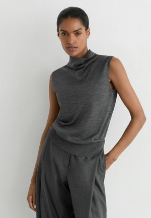 Vrouw met donker haar in een mouwloze grijze top met hoge hals en bijpassende wijde broek, staand tegen een witte muur.