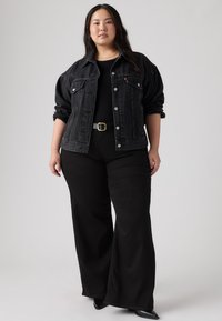 Γυναίκα που στέκεται φορώντας μαύρο τζιν μπουφάν πάνω από μαύρο τοπ, μαύρο παντελόνι με φαρδύ πόδι και ζώνη, και μαύρα παπούτσια μυτερά σε γκρίζο φόντο.