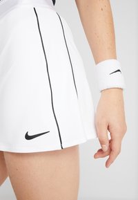 Falda de tenis blanca con rayas verticales negras y un logo de Nike negro en el dobladillo. Acompañada de una muñequera blanca con logo negro.