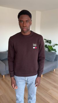 Νέος άνδρας που στέκεται μέσα σε κλειστό χώρο, φοράει ένα σκούρο καφέ φούτερ Tommy Hilfiger και ανοιχτό μπλε τζιν, με ένα γκρι καναπέ και ένα πράσινο φυτό στο φόντο.