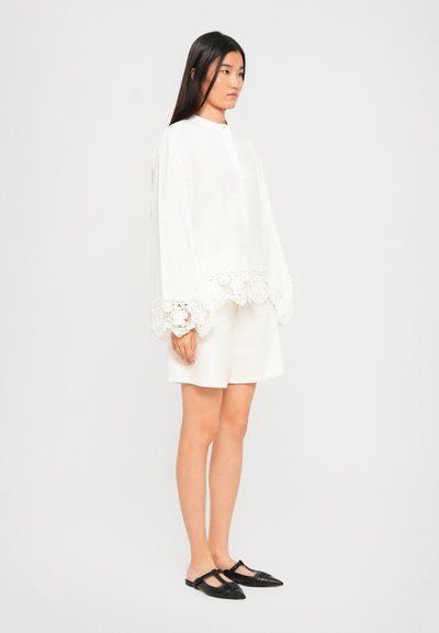 Woman standing sideways wearing a loose white blouse with lace trim, matching shorts, and black pointed flat shoes on white background.