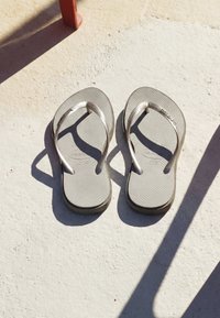 O pereche de papuci Havaianas bej, așezați unul lângă altul pe un podea texturată din beton, cu umbre de obiecte din apropiere.