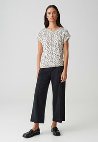 Lichte, kortste mouwen witte blouse met zwarte stippen, gecombineerd met losse zwarte broek en zwarte loafers op een neutrale achtergrond.