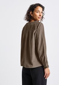 Donna con capelli ricci che indossa una blusa lunga e ampia marrone con maniche lunghe e pantaloni neri, mentre guarda oltre la spalla contro uno sfondo semplice.