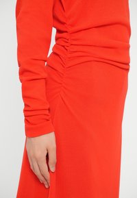 Rdeča obleka z dolgimi rokavi, ki vključuje nabran teksturo ob strani, mehko tkanino in prilegajočo zasnovo, s roko, ki počiva ob strani.