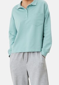 Polo de manga larga color turquesa claro con cuello, tapeta de botones y un bolsillo en el pecho, combinado con pantalones de chándal gris. Textura suave.