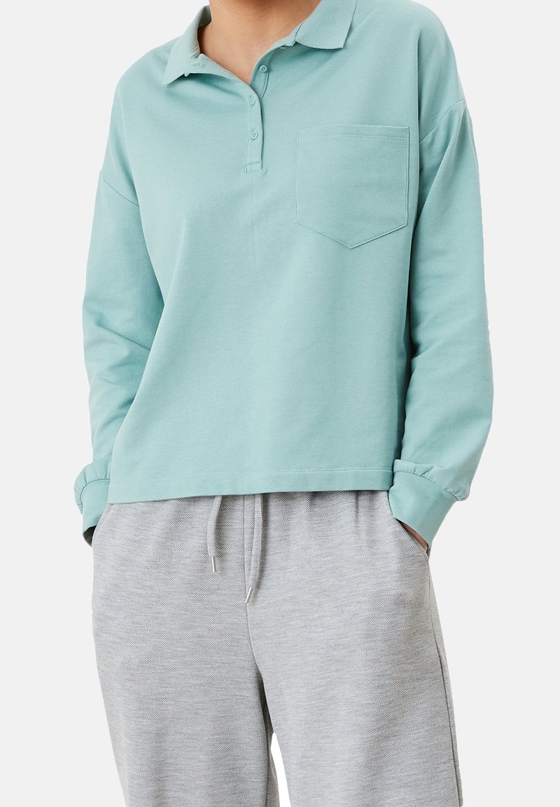 Polo de manga larga color turquesa claro con cuello, tapeta de botones y un bolsillo en el pecho, combinado con pantalones de chándal gris. Textura suave.