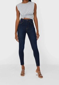 Jeans de talle alto azul oscuro con costuras naranjas, diseño ajustado y textura suave. Combinados con una blusa sin mangas gris y tacones transparentes.