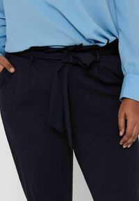 Woman wearing navy blue high-waist pants with a tied fabric belt and light blue long-sleeve blouse, hand resting in pocket.