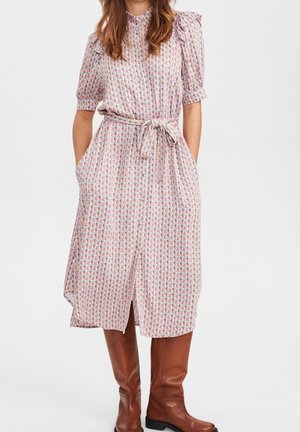 Kobieta ubrana w pastelową sukienkę midi z wzorem, z rękawami do łokci i paskiem wiązanym w talii, zestawioną z brązowymi kozakami sięgającymi do kolan.