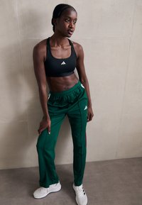 Zwart atletisch crop top met wit logo, gecombineerd met groene trackbroeken met witte accenten en een rode streep, gedragen met witte sneakers.