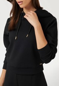 La felpa nera con cappuccio presenta un design testurizzato con un motivo sottile, cordini con punte dorate e polsini a coste.