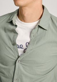 Camicia button-up verde chiaro in morbido tessuto con una vestibilità rilassata, indossata sopra una t-shirt bianca con grafica. Presenta un delicato dettaglio con logo.
