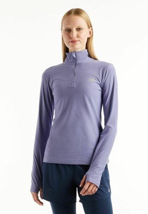 Femme portant un haut de sport à manches longues violet clair avec un col zippé et un short bleu marine, debout devant un fond uni.