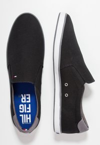 Ein Paar schwarze Slip-On-Canvas-Schuhe mit weißer Sohle, grauem Fersenakzent und blauen Einlegesohlen, bedruckt mit weißem "TOMMY HILFIGER"-Text.