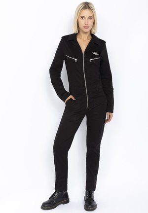Sort jumpsuit lavet af robust stof, med zip foran, to lynlåslommer på brystet, lange ærmer, tætsiddende ben og krave. Sorte støvler komplimenterer outfittet.