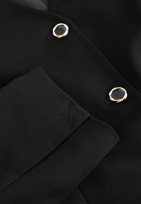 Tessuto nero su misura con un bottone con accenti dorati e pietra nera, dettaglio del logo cucito sul polsino, texture liscia.