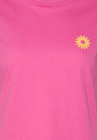 Rosa bomullströja med en gul tusensköna-grafik på vänster bröst. Mjuk textur och korta ärmar.