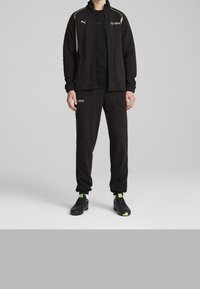 Zwart fleece trainingspak met een jack met ritssluiting, groene accenten en logo's. De broek heeft een losse pasvorm met zakken. Zwarte sneakers met groene details.