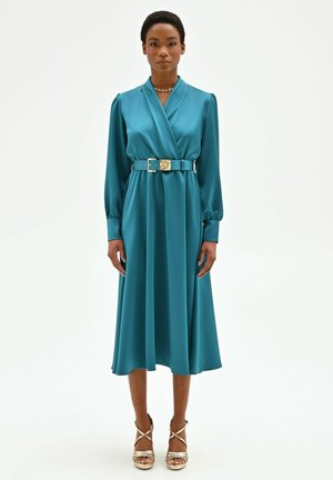 Donna in piedi che indossa un abito midi in satin turchese con maniche lunghe, una cintura larga e sandali dorati con tacco alto su uno sfondo bianco semplice.