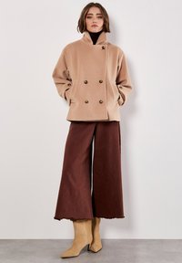 Cappotto beige doppiopetto con colletto alto, due tasche frontali, indossato su un dolcevita scuro e pantaloni marroni larghi, abbinato a stivali beige a punta.