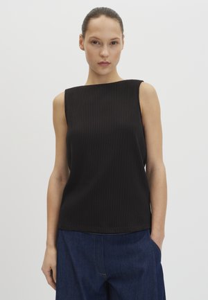 Donna con capelli tirati indietro, indossa un top nero senza maniche a coste e pantaloni larghi blu scuro, in piedi su uno sfondo chiaro uniforme.