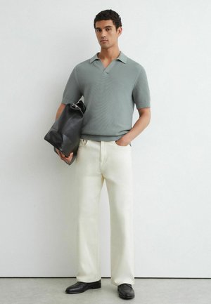 Man draagt een grijs gebreid shirt met korte mouwen, witte broek, zwarte schoenen, houdt een zwarte leren tas vast tegen een witte muur als achtergrond.