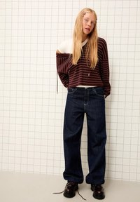 Pull court rayé bordeaux avec manches larges, associé à un jean large bleu foncé. Le mannequin porte des chaussures noires, sur un fond en carreaux.