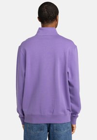 Felpa viola con colletto alto, polsini a costine e orlo, caratterizzata da una texture liscia. Modello che indossa jeans in denim blu.