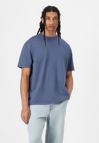 T-shirt de algodão azul com mangas curtas, corte solto, decote redondo e ligeira textura. Combinado com jeans de ganga azul clara.