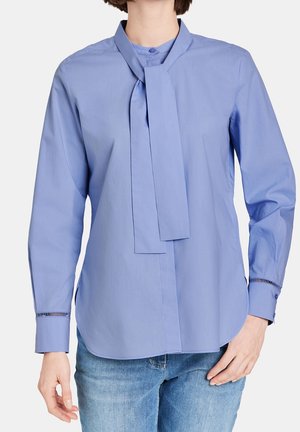 Woman wearing light blue long-sleeve blouse with neck tie detail and blue jeans, standing against a plain background.