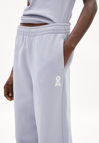 Pantalones de chándal de color púrpura claro, confeccionados en tela suave, con una cinturilla elástica, bolsillos laterales y un logo blanco bordado en la parte inferior de la pierna.