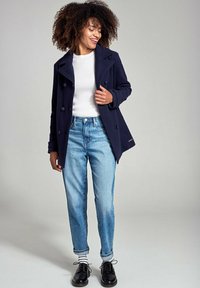 Cappotto peacoat in lana blu navy con bottoni doppi, indossato sopra un maglione bianco a coste e abbinato a jeans azzurri chiari e scarpe nere stringate.