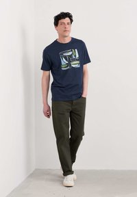 Námořnická modrá bavlněná tričko s abstraktním grafickým designem, spárované se světlými olivově zelenými kalhotami a béžovými teniskami. Model stojí proti světlé zdi.