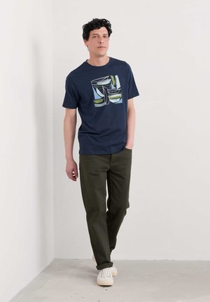 T-shirt en coton bleu marine avec un graphique abstrait, associé à un pantalon vert olive et des baskets beiges. Modèle se tenant contre un mur clair.