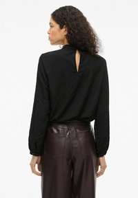 Svart teksturert bluse med n�økkelhull-detalj bak, kombinert med burgunder høytlivs skinnbukser med to baklommer.