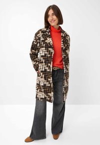 Lange, gestreepte jas in bruin, Room en zwart met een ingesneden kraag, gecombineerd met een rode trui, grijze uitlopende broek en beige schoenen.