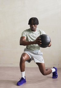Atlet v lahkorozeleni majici Nike in sivih kratkih hlačah izvaja izpad s črno medicinsko žogo na nevtralni podlagi.