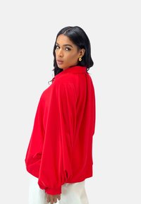 Red oversized long-sleeve shirt with a collar, featuring gathered sleeves and a smooth texture, paired with loose white pants.