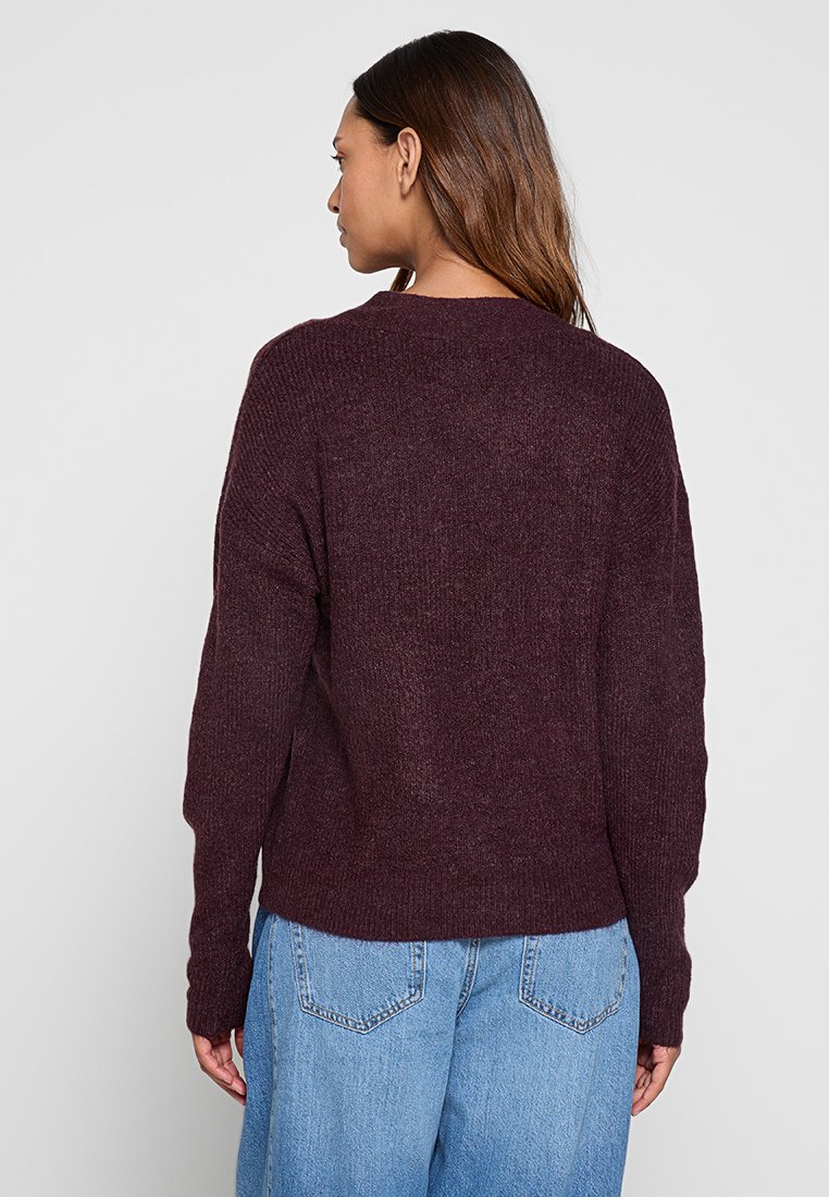 Bordeauxroter Strickpullover mit lockerer Passform, Rundhalsausschnitt, überschnittenen Schultern und gerippten Bündchen, kombiniert mit hellblauen Jeans.