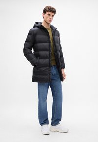 Chaqueta negra acolchada con capucha, cierre de cremallera y bolsillos laterales; combinada con camisa verde y jeans azules, llevada con zapatillas blancas.