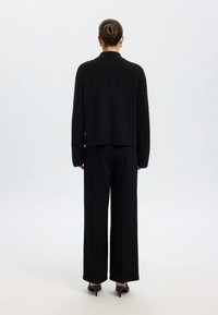 Juodas, minkštas, tekstūruotas megztinis su aukštu kaklu, derinamas su platėjančių kojų juodomis kelnėmis ir stilingais juodais aukštakulniais batais.