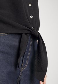 Černá blůza s texturovaným povrchem, s ozdobnými stříbrnými knoflíky, kombinovaná s modrými denimovými džíny, které mají kontrastní žluté švy.