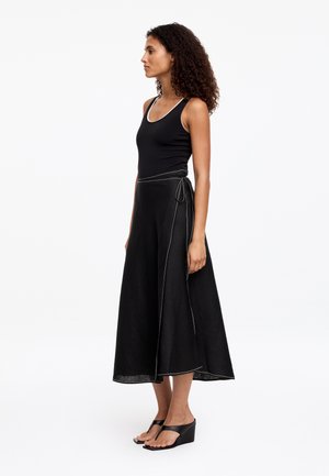 Kobieta o kręconych włosach, ubrana w czarny top bez rękawów, czarną midi spódnicę kopertową z białymi szwami oraz czarne sandały na koturnie, stojąca bokiem.