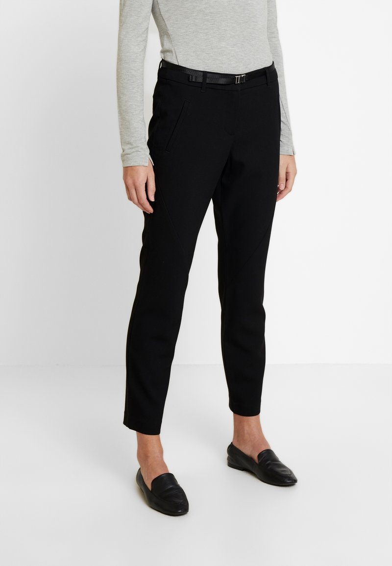Pantalon noir sur mesure avec une texture lisse, doté de poches latérales et d'une coupe slim, associé à un haut gris à manches longues et des chaussures noires sans lacets.