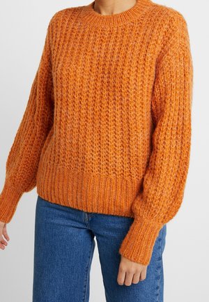 Personne portant un pull orange épais en tricot texturé à manches longues et un jean bleu sur un fond clair uni.